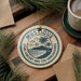 A circular ornament with the text "MUSTIQUE SAINT VINCENT AND THE GRENAOINES" printed on it, surrounded by pine branches and a cup of coffee on a wooden surface.