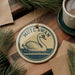A decorative ornament featuring a swan, with the text "MUTE SWAN" printed on it, is displayed on a wooden surface alongside a cup of coffee and some pine branches.
