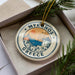 A decorative ornament in a box, featuring a circular design with a depiction of a Greek island and a windmill, surrounded by greenery.