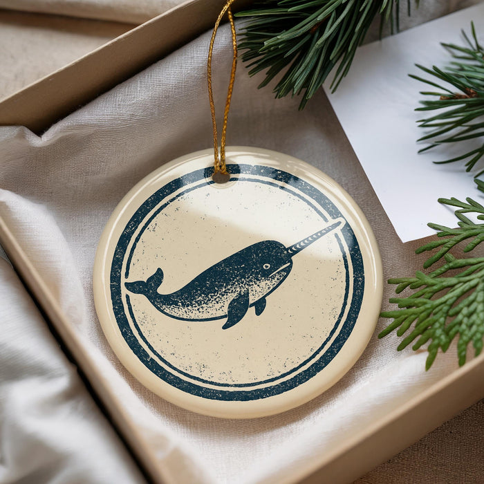 A round ornament featuring a narwhal is displayed in a box, surrounded by pine branches.