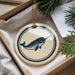 A round ornament featuring a narwhal is displayed in a box, surrounded by pine branches.