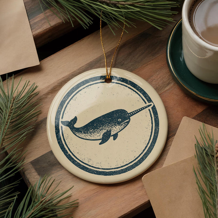 a circular ornament with a nautical design, including a whale and a harp, hanging on a wooden surface. There is also a cup of coffee and some pine branches in the background.
