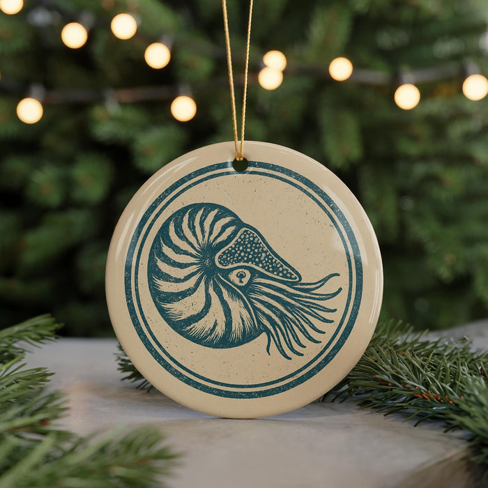 A circular ornament featuring a nautilus shell design, hanging from a string and placed on a surface with pine branches in the background.
