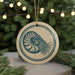 A circular ornament featuring a nautilus shell design, hanging from a string and placed on a surface with pine branches in the background.