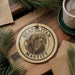 A circular ornament featuring a dog's face and the text "NEAPOLITAN MASTIFF" is displayed on a wooden surface, surrounded by pine branches and a cup of coffee.