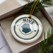 A white ceramic ornament with the word "NIUE" in blue text, surrounded by pine branches and greenery.