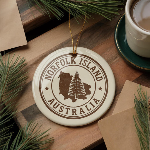 A round ornament with the text "NORFOLK ISLAND AUSTRALIA" and a tree silhouette is displayed on a wooden surface, surrounded by pine branches and a cup of coffee.