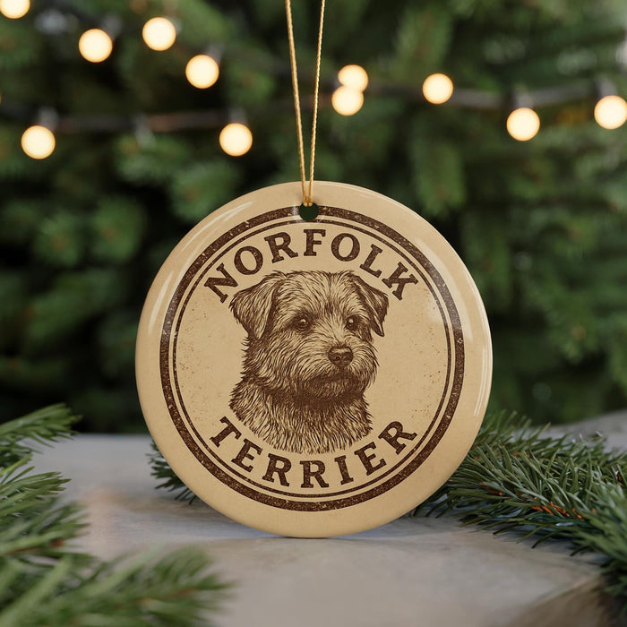 A round ornament featuring a dog's head and the text "NORFOLK TERRIER" is displayed in front of a blurred Christmas tree.