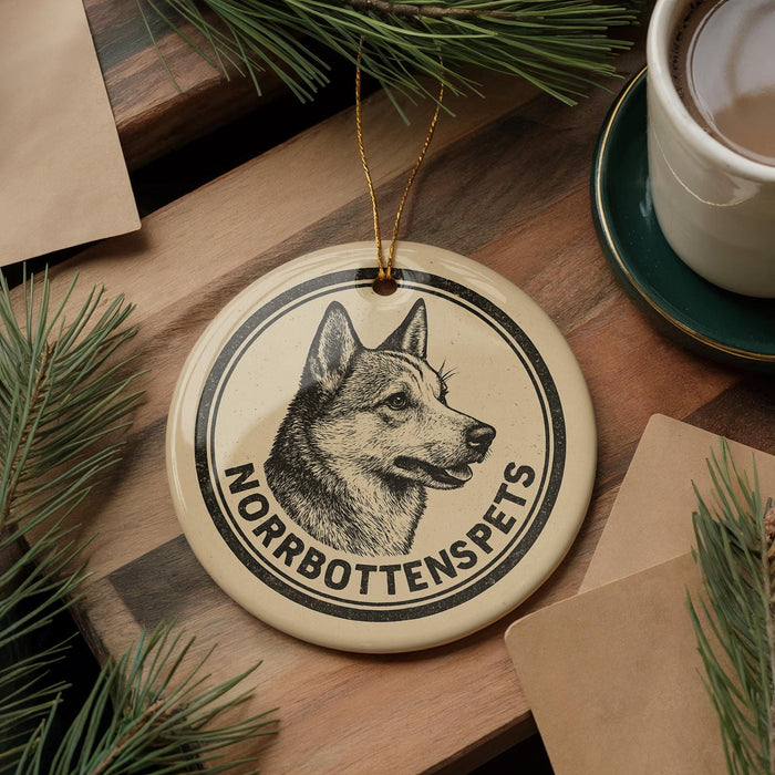 A round ornament featuring a black and white illustration of a dog's head, with the text "NORROBOTTENSPETS" printed on it, is displayed on a wooden surface alongside a cup of coffee and some pine branches.
