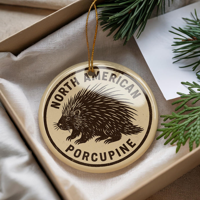 A porcupine ornament is displayed in a box, surrounded by pine branches and a white card.