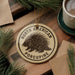 A porcupine ornament with the text "NORTH AMERICAN PORCUPINE" is displayed on a wooden surface, surrounded by pine branches and a cup of coffee.