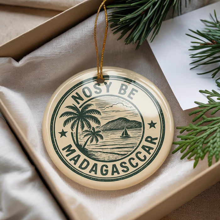 A circular ornament with the text "NOSY BE MADAGASCAR" printed on it, surrounded by a palm tree and a sailboat, is displayed in a box with pine branches.