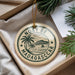 A circular ornament with the text "NOSY BE MADAGASCAR" printed on it, surrounded by a palm tree and a sailboat, is displayed in a box with pine branches.