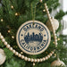 A circular ornament on a Christmas tree with the text "OAKLAND CALIFORNIA U.S.A." printed on it, surrounded by other ornaments and lights.