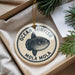 A round ornament with a fish design, hanging from a string, placed inside a box with pine branches.