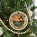 A circular ornament on a Christmas tree, featuring a palm tree and a sunset landscape, with the text "OKAVANGO DELTA BOTSWANA" around the edge.