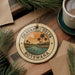 A circular ornament with the text "OKAVANGO DELTA Botswana" and a tropical landscape design, placed on a wooden surface next to a cup of coffee and some pine branches.