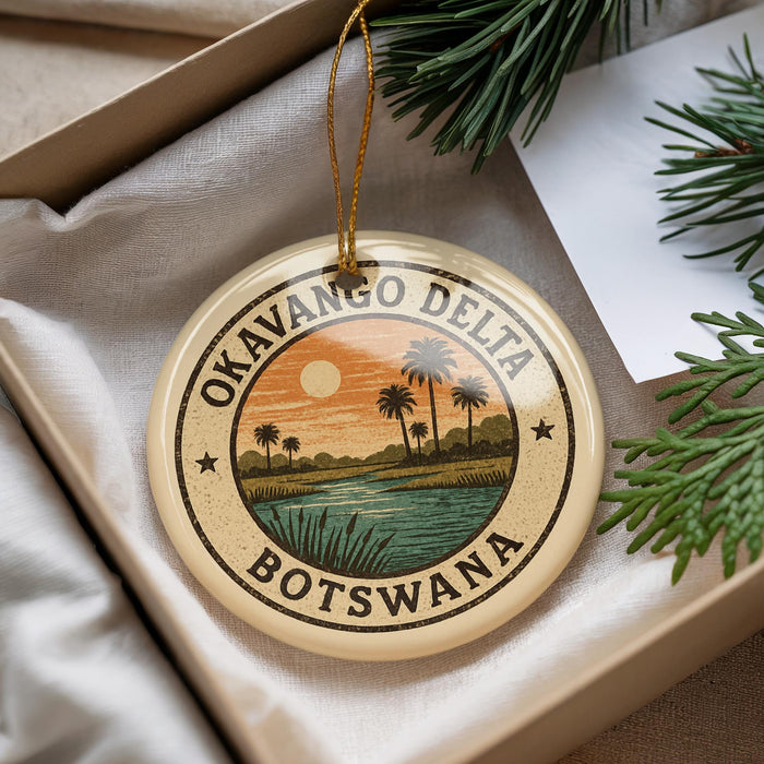A circular ornament with a tropical landscape design, featuring palm trees, a sunset, and a body of water, is displayed in a box with pine branches.