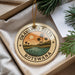 A circular ornament with a tropical landscape design, featuring palm trees, a sunset, and a body of water, is displayed in a box with pine branches.