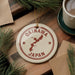 A circular ornament with the text "OKINAWA JAPAN" printed on it, surrounded by pine branches and a cup of coffee on a wooden surface.