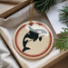 A circular ornament featuring a black and white orca whale silhouette is displayed in a box, surrounded by pine branches.