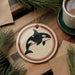 a circular ornament with a black and white orca (or killer whale) silhouette, hanging on a wooden surface. The ornament is surrounded by pine branches and a cup of coffee, creating a cozy and festive atmosphere.