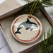 A ceramic ornament in the shape of a black and white orca whale, with a red border, is displayed in a box with pine branches.