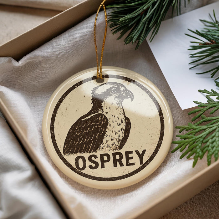 A circular ornament with a bird image and the word "OSPREY" printed on it, placed on a surface with pine branches and a white paper.