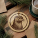 a round ornament with a brown and white illustration of an otter on a wooden surface, surrounded by pine branches and a cup of coffee.