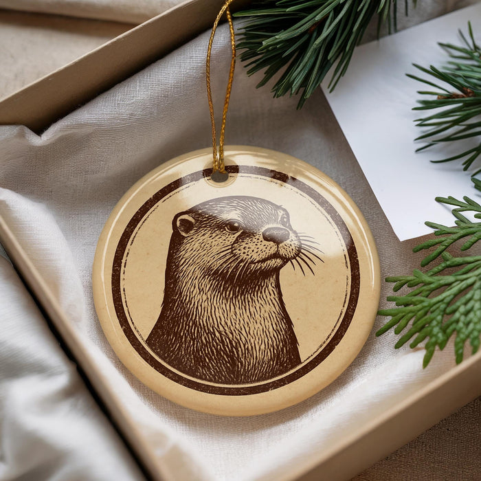 A round ornament featuring an image of an otter is displayed in a box, surrounded by pine branches.