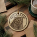 A circular ornament featuring a cat's image, with the text "PALLAS'S CAT" printed on it, is displayed on a wooden surface alongside a cup of coffee and some pine branches.