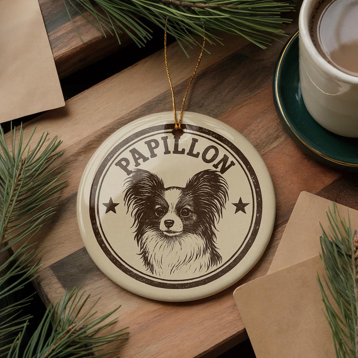 A circular ornament featuring a Papillon dog's face, with the word "Papillon" written on it, is displayed on a wooden surface alongside a cup of coffee and some pine branches.