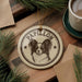 A circular ornament featuring a Papillon dog's face, with the word "Papillon" written on it, is displayed on a wooden surface alongside a cup of coffee and some pine branches.