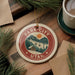 A circular ornament with the text "PARK CITY UTAH" and a mountain landscape design, hanging on a wooden surface next to a cup of coffee and pine branches.