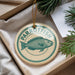 A round ornament with a parrotfish design, hanging from a string, placed inside a box with pine branches.