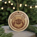 A circular ornament featuring a Pekingese dog's face, with the text "Pekingese Dog" printed on it, is displayed in front of a blurred Christmas tree.