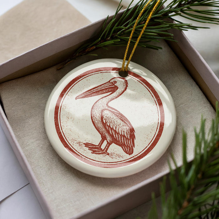 A white ceramic ornament with a red and brown illustration of a pelican is displayed in a box, surrounded by pine branches.