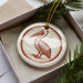 A white ceramic ornament with a red and brown illustration of a pelican is displayed in a box, surrounded by pine branches.