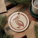 a round ornament with a pelican design, placed on a wooden surface next to a cup of coffee and some pine branches.