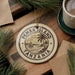 A circular ornament with the text "PEMBA ISLAND TANZANIA" and a graphic of a sailboat on the water, surrounded by pine branches and a cup of coffee on a wooden surface.