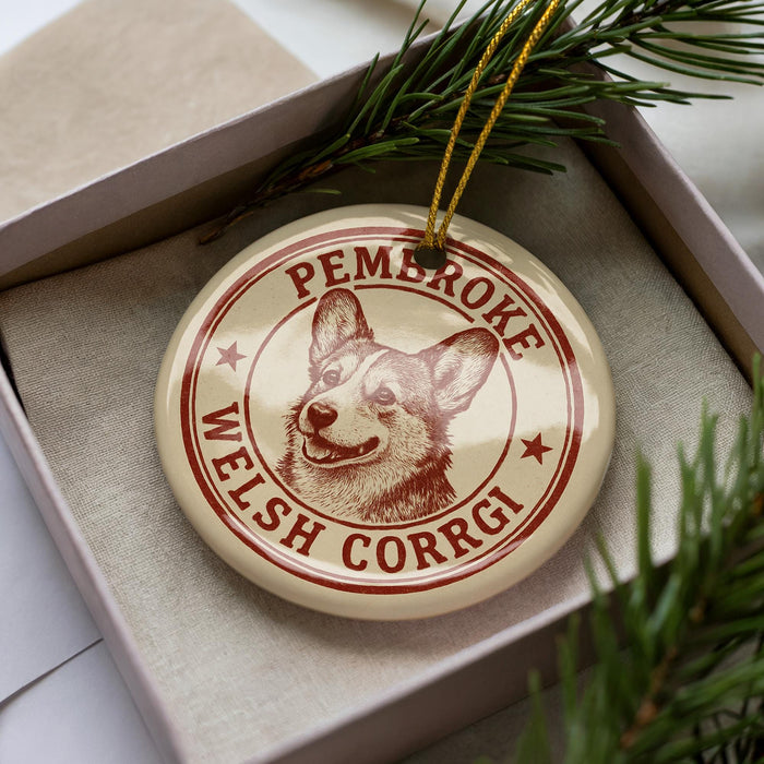 A circular ornament with a Welsh Corgi dog image and the text "PEMBROKE WELSH CORRGI" printed on it, placed inside a box with pine branches.