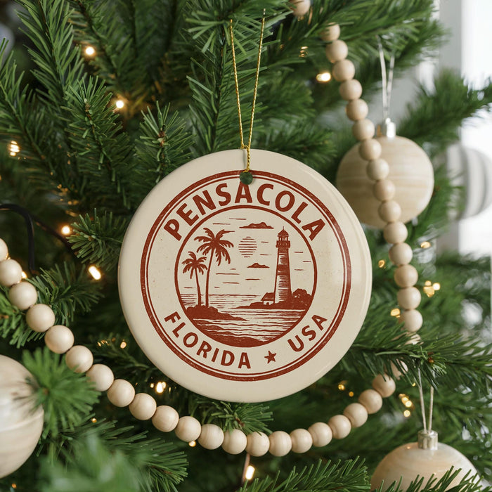 A round ornament on a Christmas tree, featuring a lighthouse and palm trees, with the text "Pensacola, Florida USA" printed on it.
