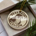 A circular ornament with the text "Peregrine Falcon" printed on it, surrounded by pine branches.