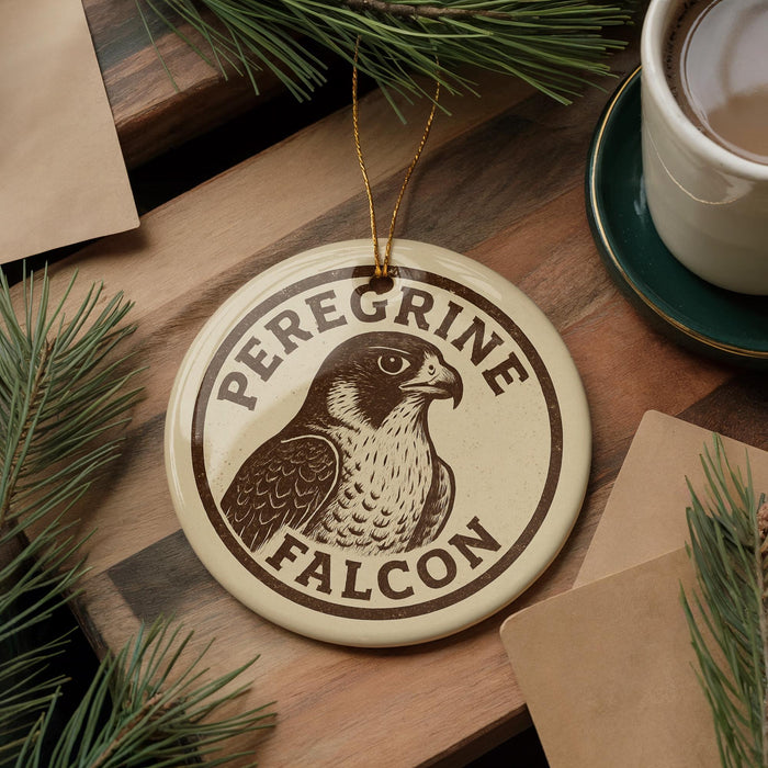 A circular ornament featuring a peregrine falcon, with the text "Peregrine Falcon" printed on it, is displayed on a wooden surface alongside a cup of coffee and some pine branches.