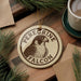 A circular ornament featuring a peregrine falcon, with the text "Peregrine Falcon" printed on it, is displayed on a wooden surface alongside a cup of coffee and some pine branches.