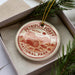 A circular ornament with the text "PETRIFIET FOREST NATIONAL PARK USA" printed on it, placed inside a box with pine branches.