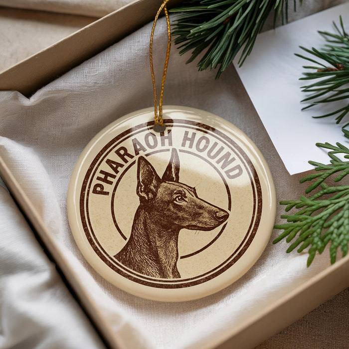 A round ornament with a dog image and the text "PHARAO HOUND" printed on it, placed in a box with pine branches.