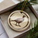 A ceramic ornament in the shape of a bird, possibly a pheasant, is displayed in a box with pine branches.