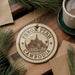 A circular ornament with the text "PHNOM PENH CAMBODIA" and an illustration of a temple, placed on a wooden surface next to a cup of coffee and some pine branches.