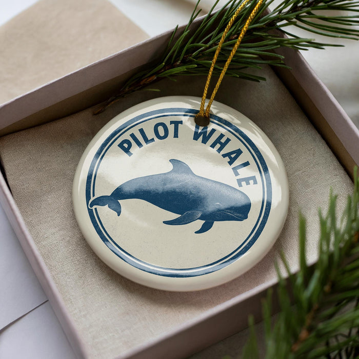 A round ornament with a blue and white design featuring a pilot whale, placed in a box with pine branches.